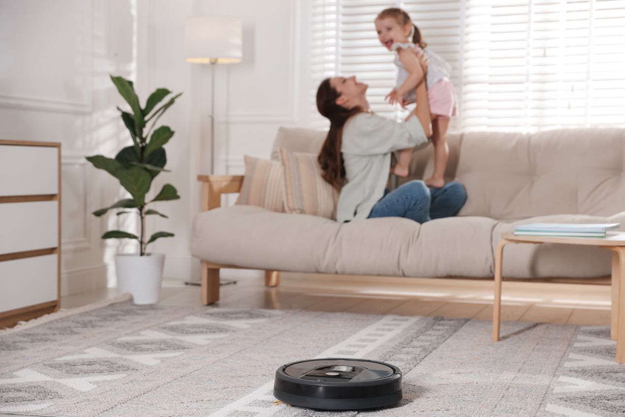 Saugroboter reinigt Teppich, waehrend Mutter mit Kind auf Sofa spielt – Symbolbild fuer strukturierte Entlastung durch Haushaelterin